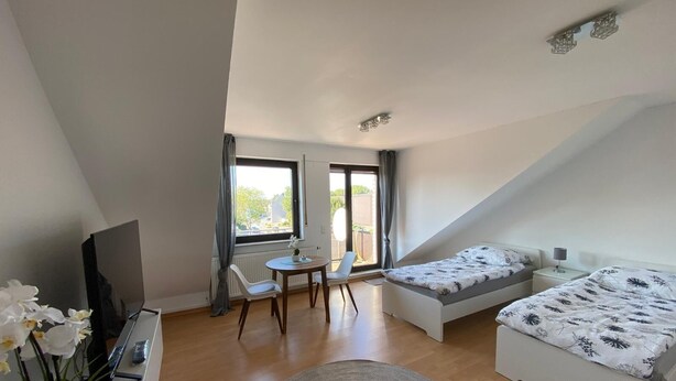 A modern, well-lit room with a bed, a desk, and a chair. There is a window providing natural light and a view of the outdoors. - Am Quellberg Apartments Recklinghausen (Recklinghausen)