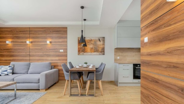 A modern living room with a grey sofa, a wooden dining table with chairs, and a kitchen area with wooden cabinetry.