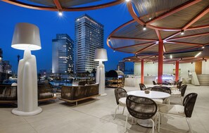 Un espacio para comer al aire libre con mesas y sillas redondas, una lámpara blanca grande y vistas de edificios altos al anochecer.
