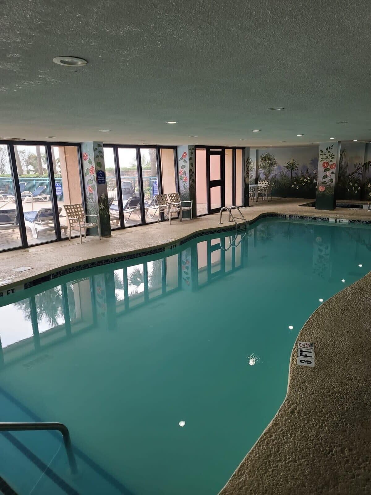 An indoor swimming pool with clear glass walls, lounge chairs, and a mural of flowers on the wall.