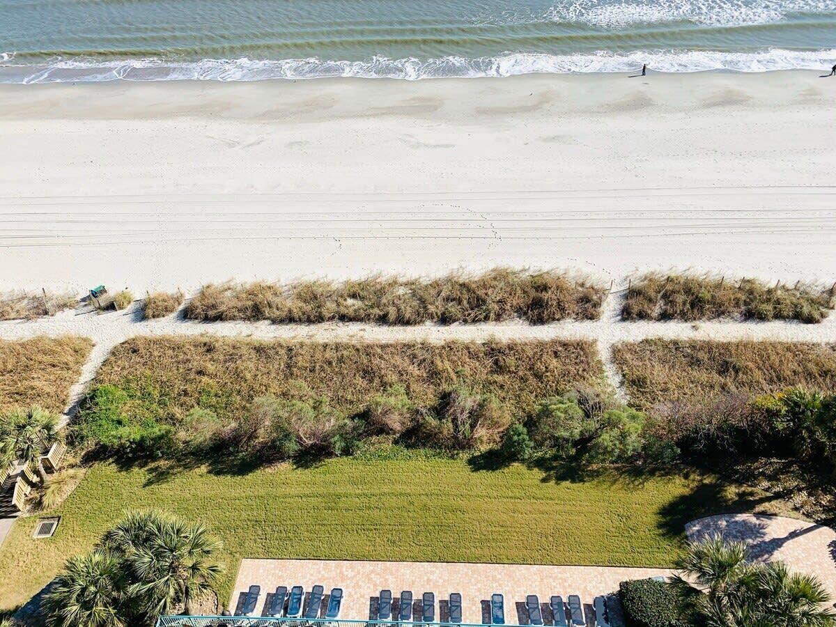 An aerial view of a beach with a sandy shore, a grassy area with pathways, and a row of blue chairs in the foreground.