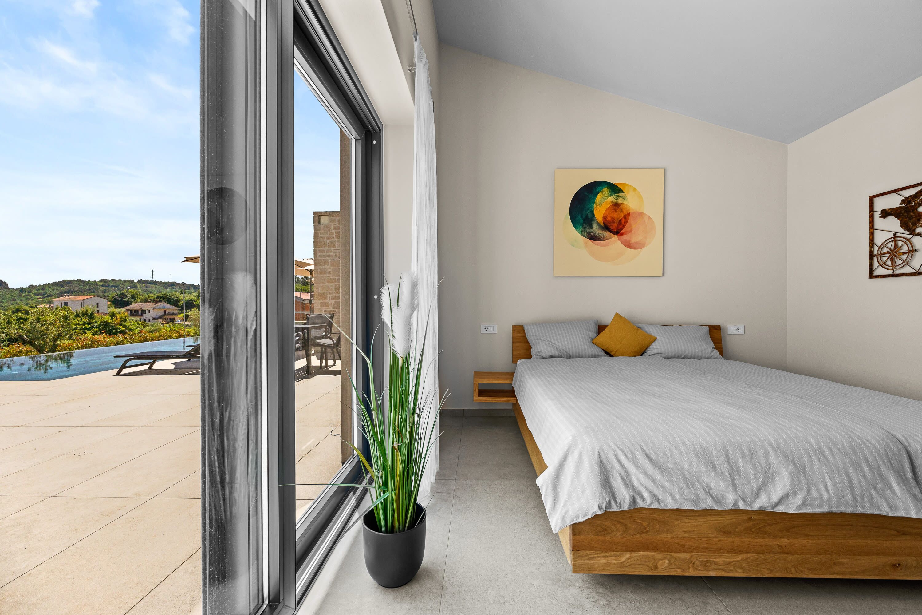 A bedroom with a large window, a bed, a potted plant, and a painting on the wall.