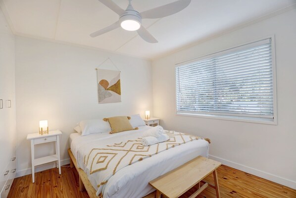 Ein Schlafzimmer mit einem Bett, Nachttischen, einem Deckenventilator, einem Fenster mit Jalousien und einem Wandbild.