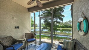 Una habitación con un amplio ventanal corredizo que da a un estanque, mobiliario de mimbre y un ventilador de techo.