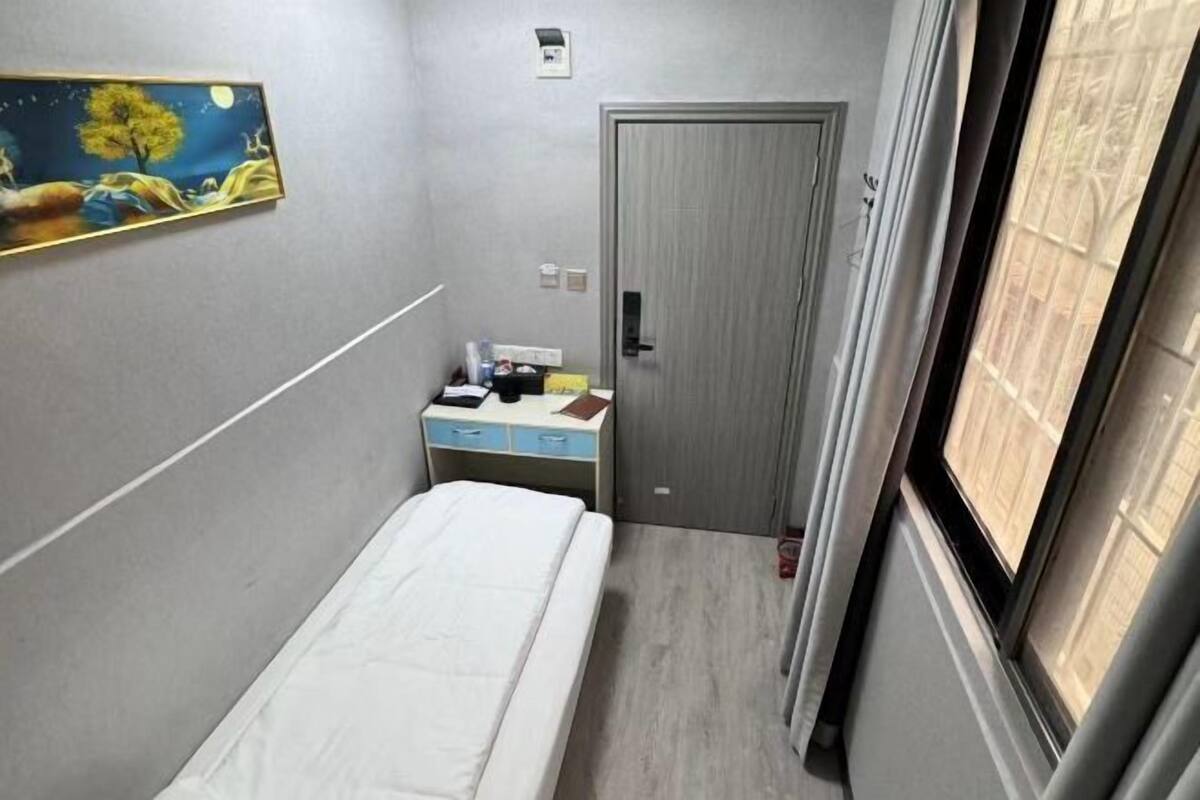 A small, narrow hotel room with a bed, a desk with a chair, a door, and a window.