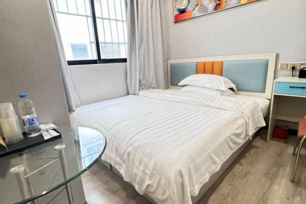 A hotel room with a bed, a glass table, a chair, a desk, a window with curtains, and a painting on the wall.