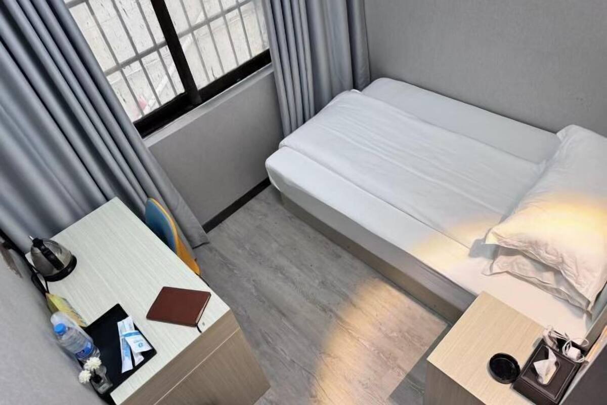 A single bed with a white mattress and pillow, a bedside table with a lamp, a small desk with a chair, and a window with curtains.