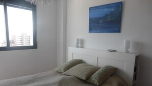Une chambre à coucher avec un lit, deux oreillers, une table de chevet, un tableau et une fenêtre donnant sur des immeubles.