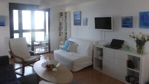 Un salon avec un canapé blanc, une table basse sur laquelle on a posé des fruits, une bibliothèque, un téléviseur et une fenêtre donnant sur un beau paysage.