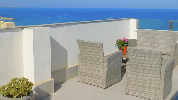 Une terrasse meublée de mobilier en osier, agrémentée d’une plante en pot, qui offre une vue sur une mer d’un bleu limpide.