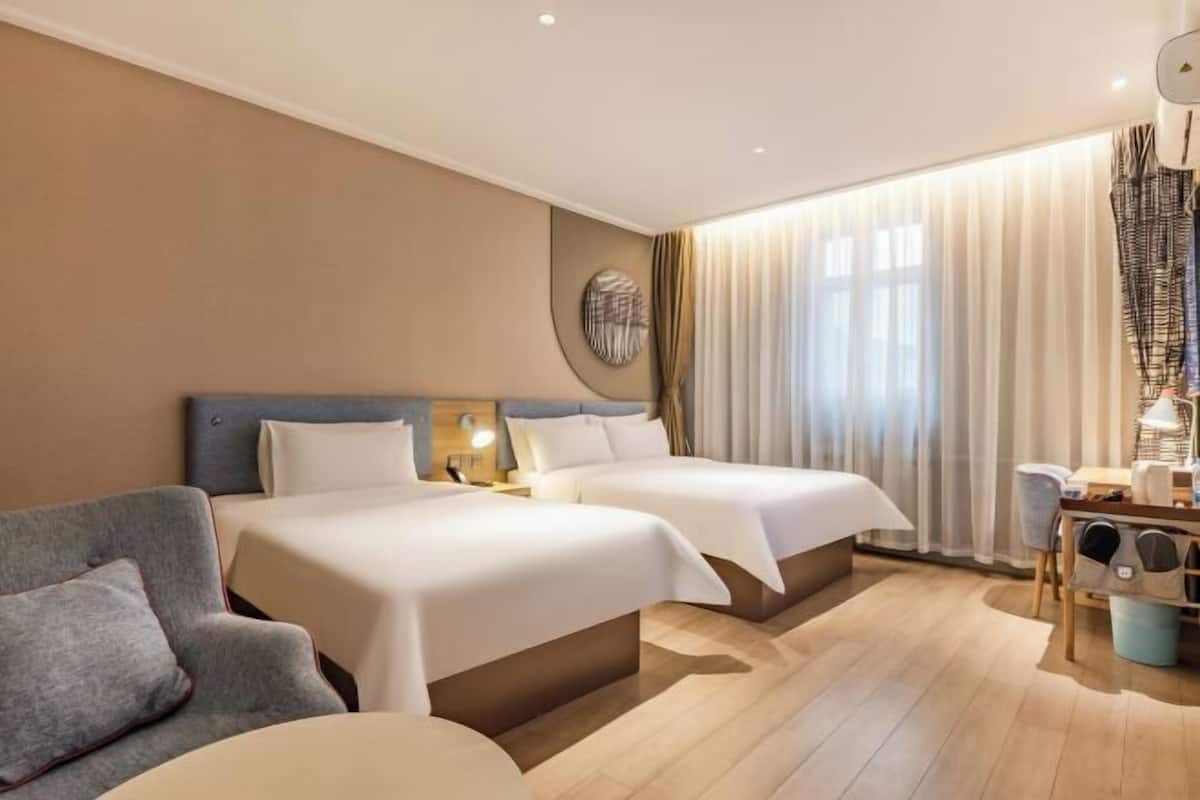 A hotel room with two beds, a sofa, a desk with a lamp, and a large window with curtains.
