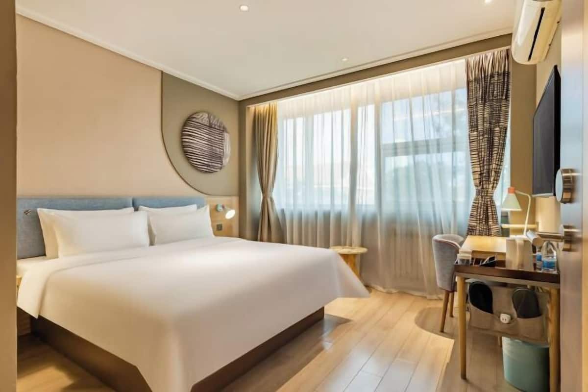 A hotel room with a bed, a desk, a chair, a lamp, and a large window with curtains.