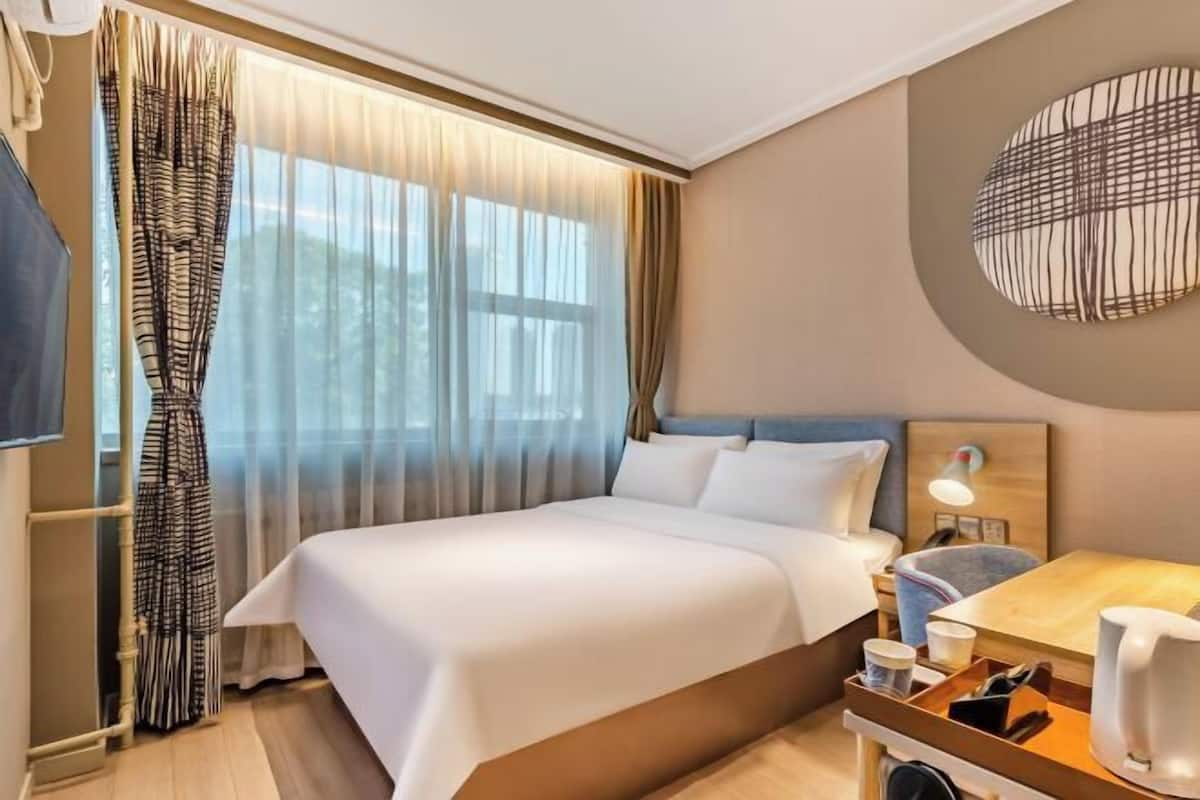 A hotel room with a bed, a bedside table with a lamp, a wall-mounted artwork, and a window with curtains.