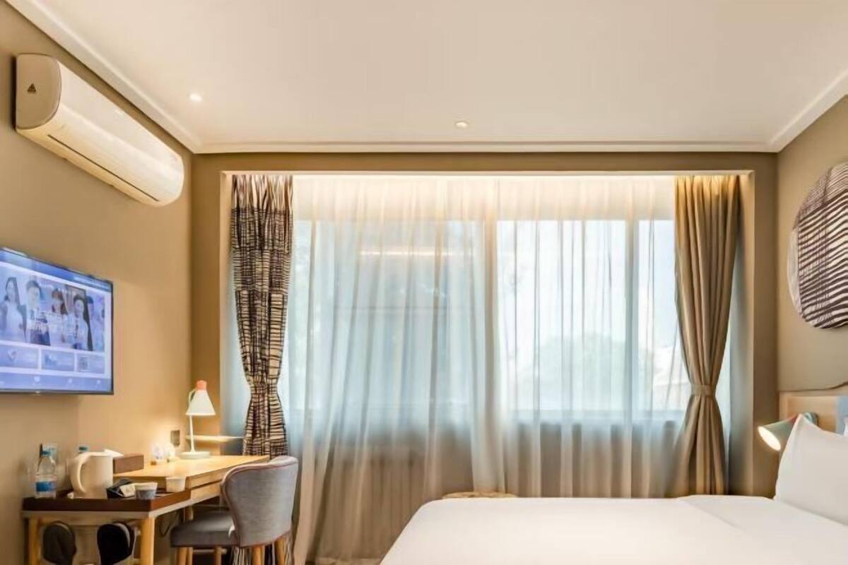 A hotel room with a bed, a desk with a chair, a television, and a window with curtains.