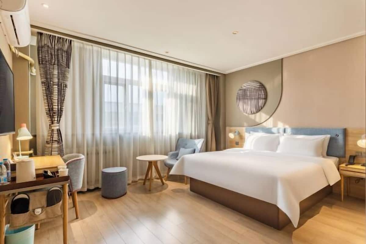 A hotel room with a bed, a desk with a chair, a round side table, a lamp, and a window with curtains.