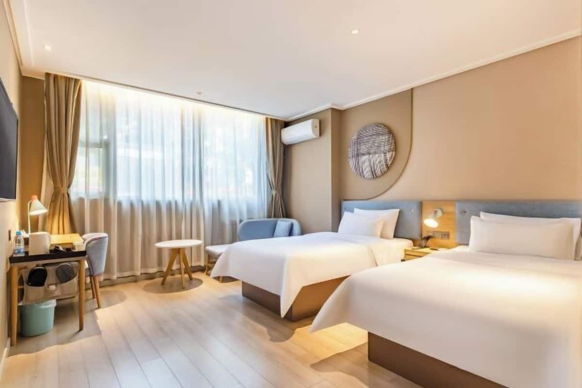 A hotel room with two beds, a desk with a chair, a small table, and a large window with curtains.