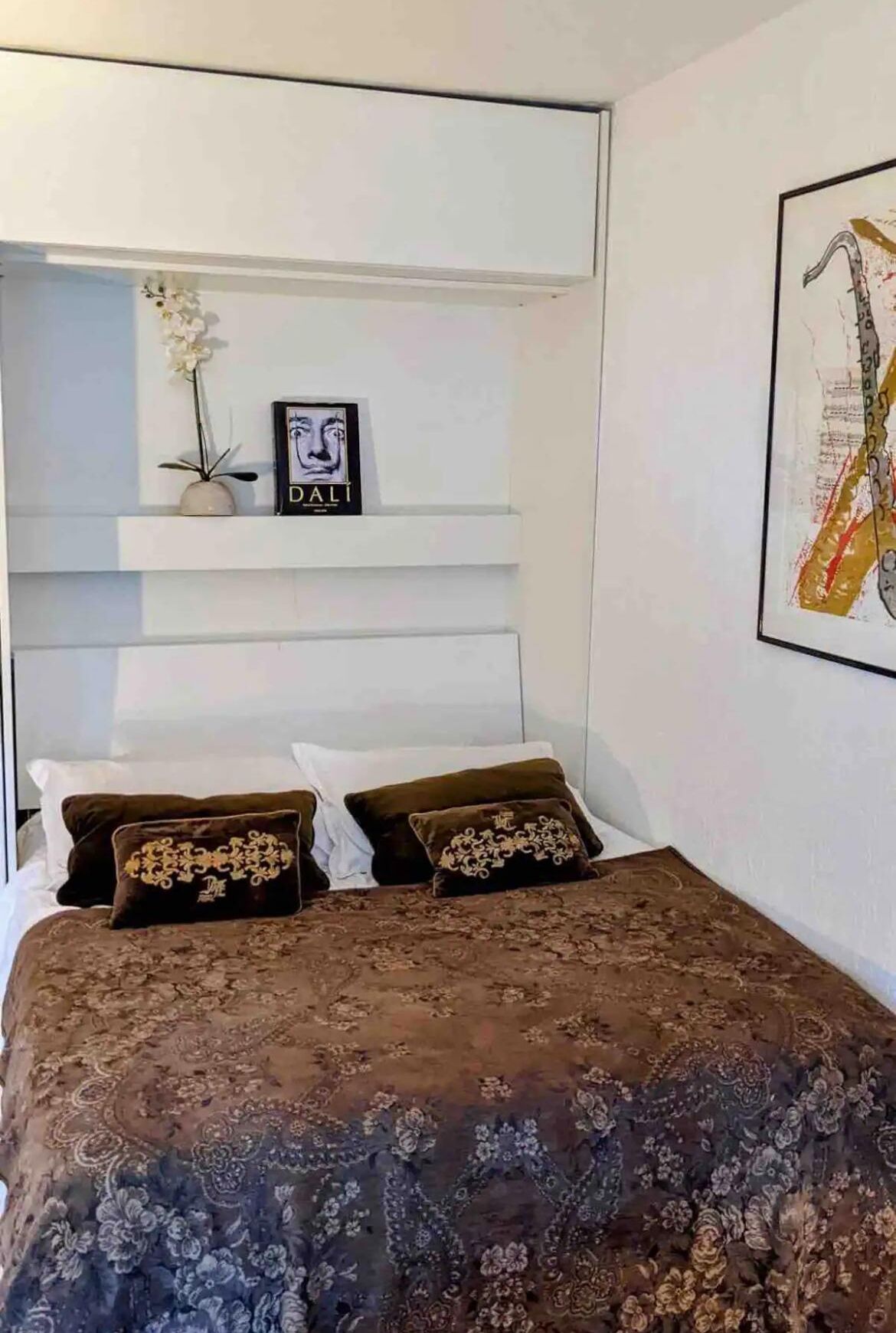 Un dormitorio con una cama, una pared blanca, un cuadro enmarcado y una estantería con un jarrón y una fotografía.