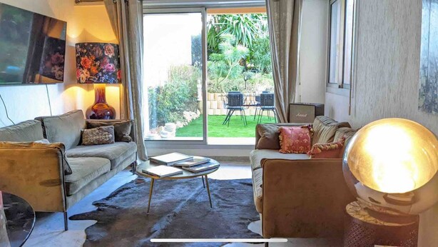 Een woonkamer met twee banken, een salontafel en een lamp. Er is een groot raam met uitzicht op een tuin en tuinmeubilair.