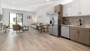 Une cuisine moderne avec des meubles en bois, un réfrigérateur en acier inoxydable et un lave-vaisselle. On y trouve un coin repas avec une table et des chaises, ainsi qu’un canapé à l’arrière-plan.
