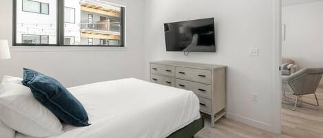 A bedroom with a bed, a TV mounted on the wall, a dresser, and a window with a view of a building.