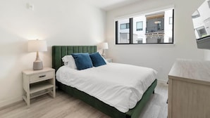 Une chambre à coucher moderne avec une tête de lit recouverte d’un tissu vert, une literie blanche et une table de chevet avec une lampe.