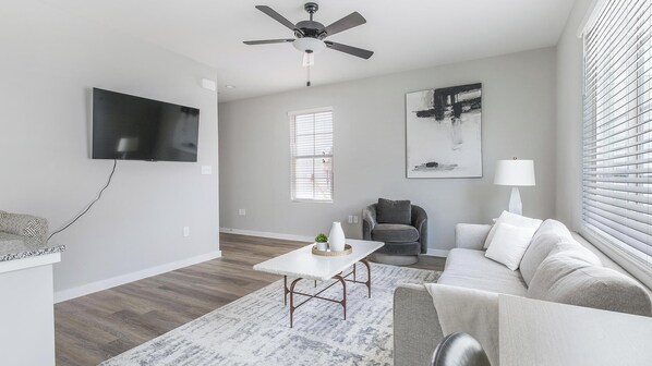 A modern living room with a flat-screen TV, a ceiling fan, a gray sofa, a coffee table, and a painting on the wall. - Landing | Stylish 1BD, Gym, Pool (Madison)