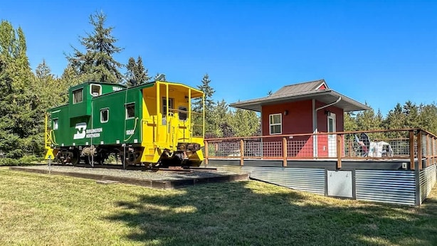 Un vagón de tren verde y amarillo con la inscripción 'Burlington Northern', un edificio rojo con cerca metálica y una zona arbolada al fondo.