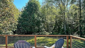 Dos sillas Adirondack en una terraza de madera con barandilla metálica, con vistas a una zona ajardinada con árboles.