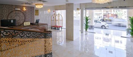 A hotel reception area with a reception desk, a chandelier, and a view of the street outside.