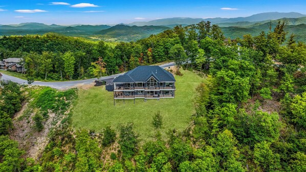 A large, two-story house with a dark roof, surrounded by dense forest and mountains in the background.