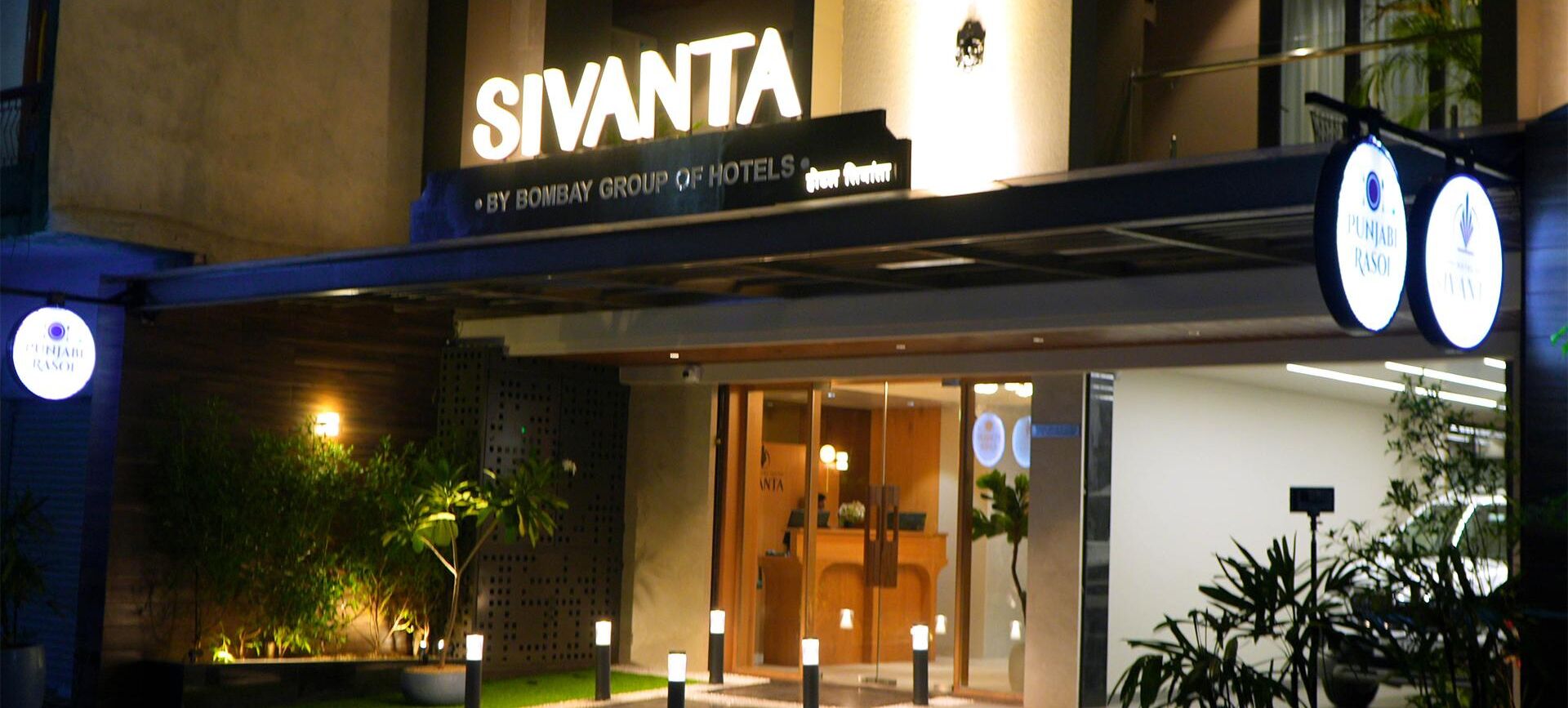 The exterior of the Sivanta hotel, featuring a prominent sign, a well-lit entrance, and a landscaped area with potted plants.