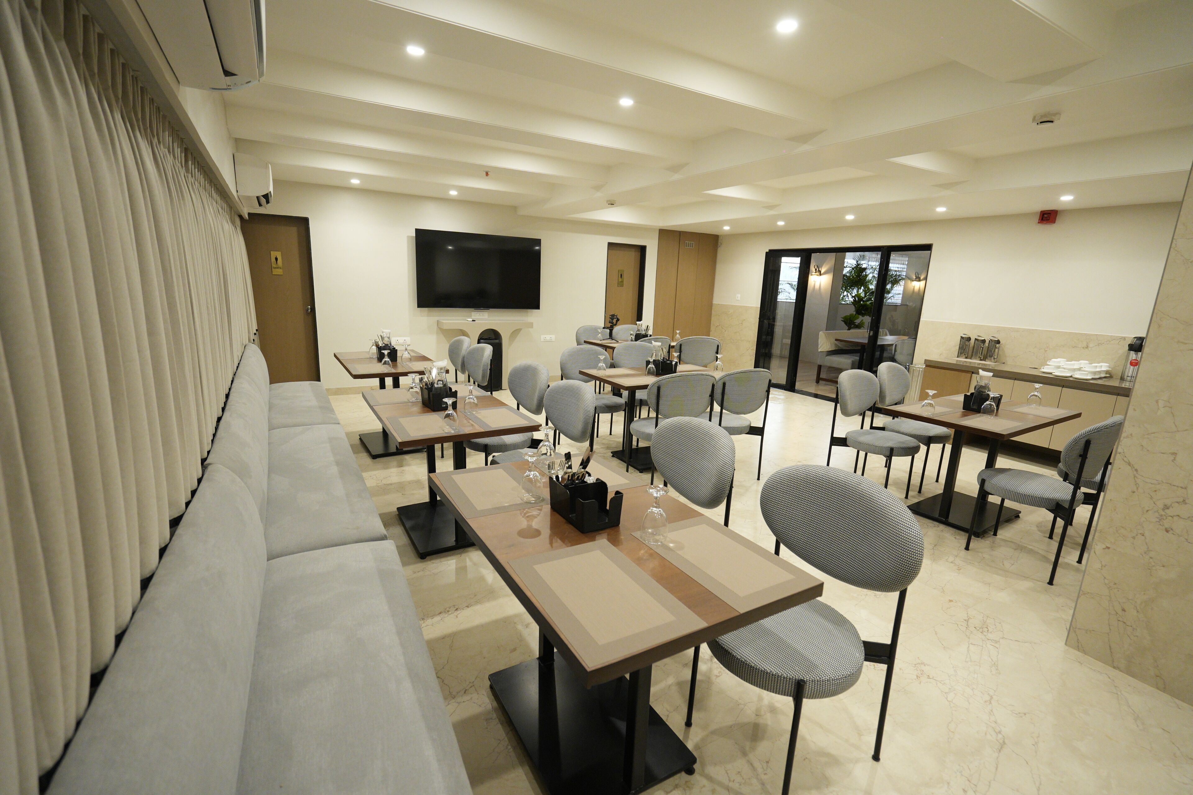 A hotel dining area with multiple tables and chairs, a flat-screen TV mounted on the wall, and a large window providing natural light.