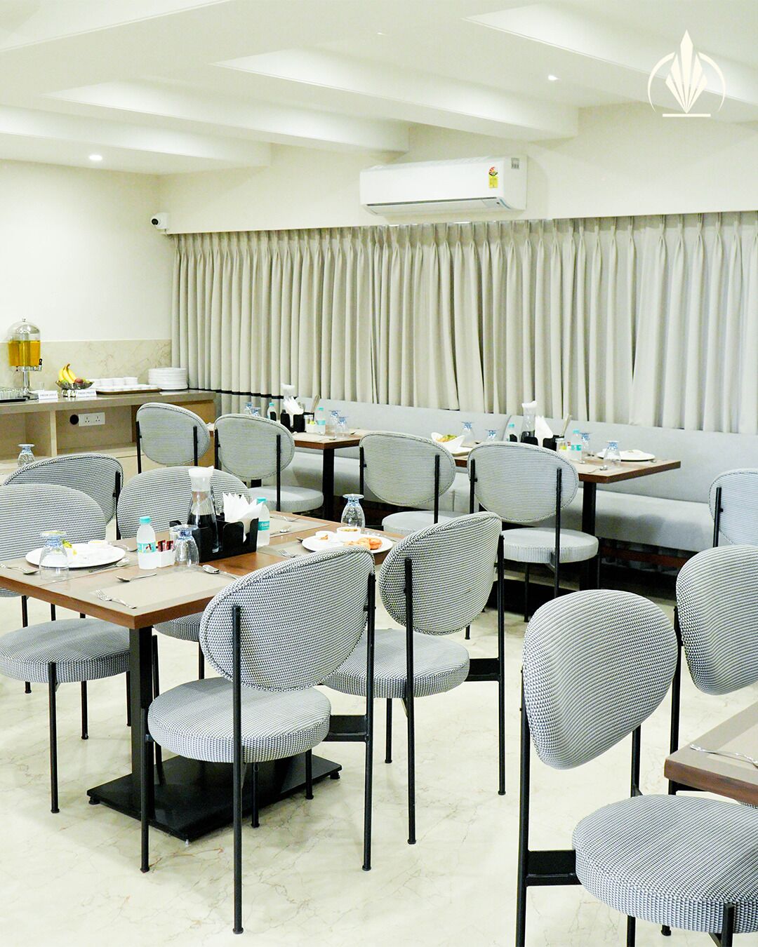 A hotel dining area with multiple tables and chairs, a buffet setup, and an air conditioning unit on the ceiling.