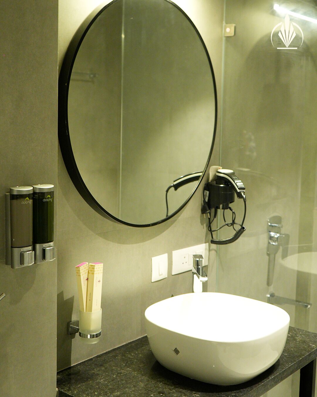A modern bathroom with a round mirror, a white sink, and a towel dispenser.