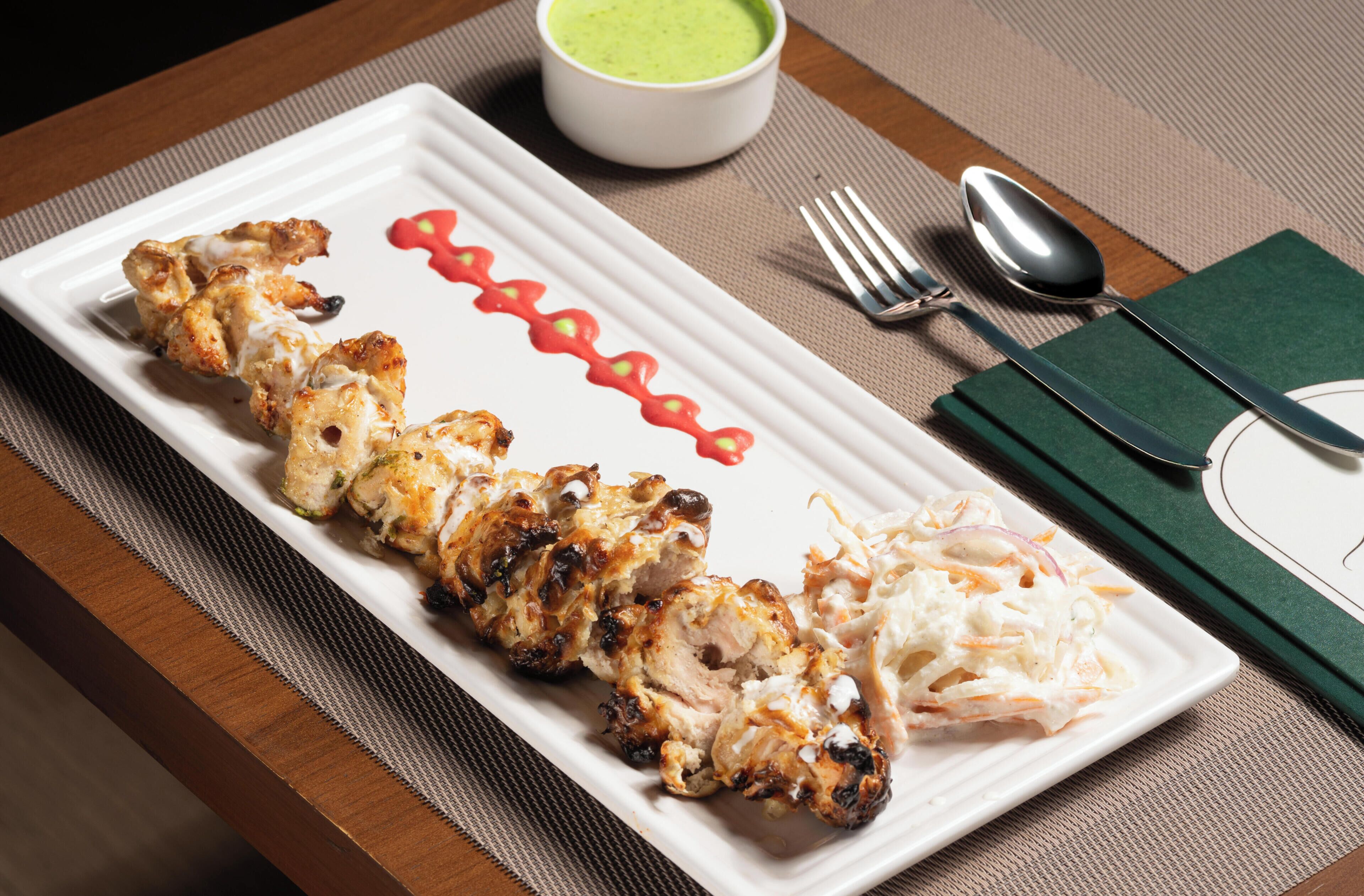 A white rectangular plate with grilled chicken skewers, coleslaw, and a green sauce on a wooden table.