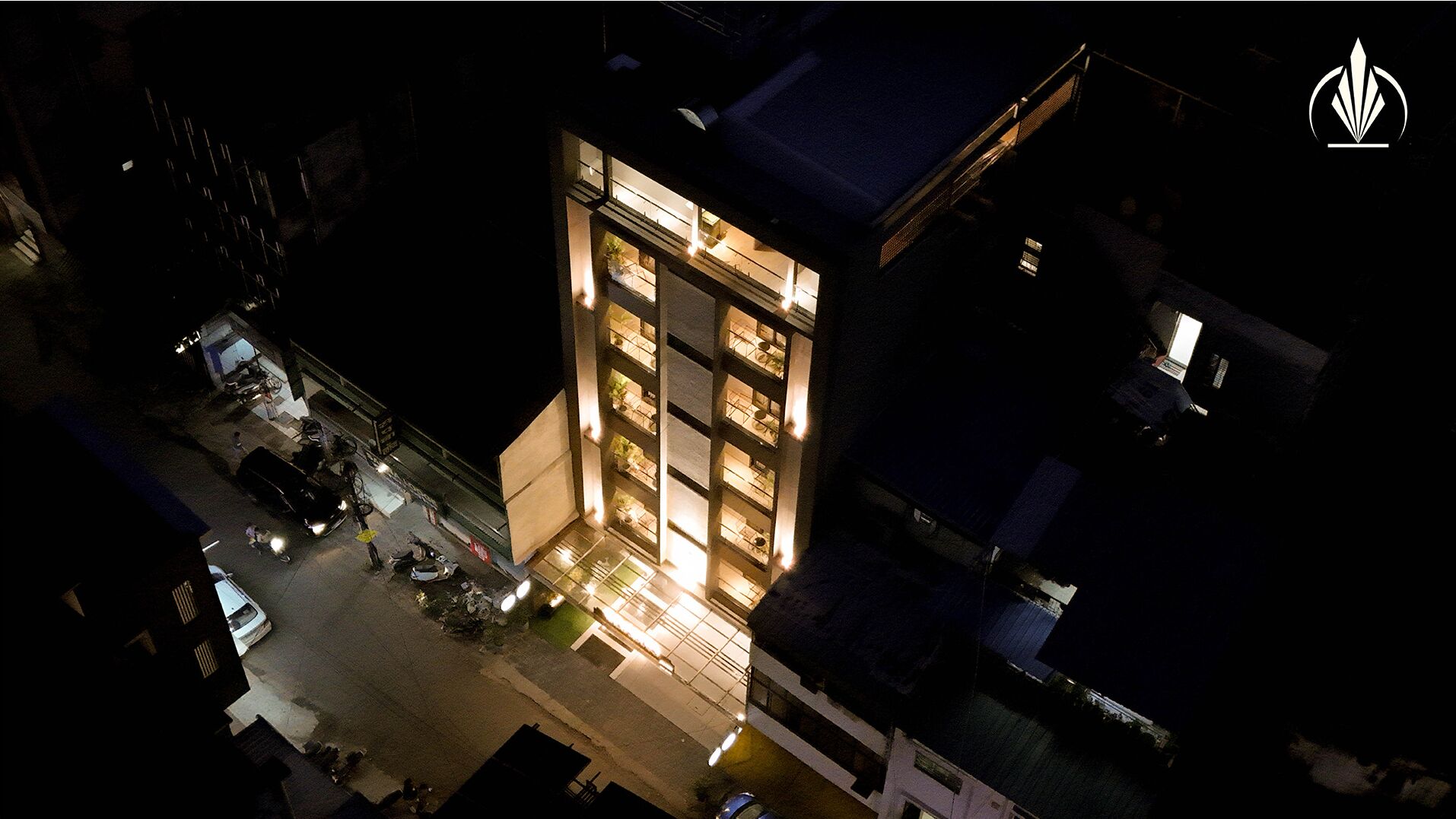A multi-story building with illuminated windows, a flat roof, and a parking area in front.