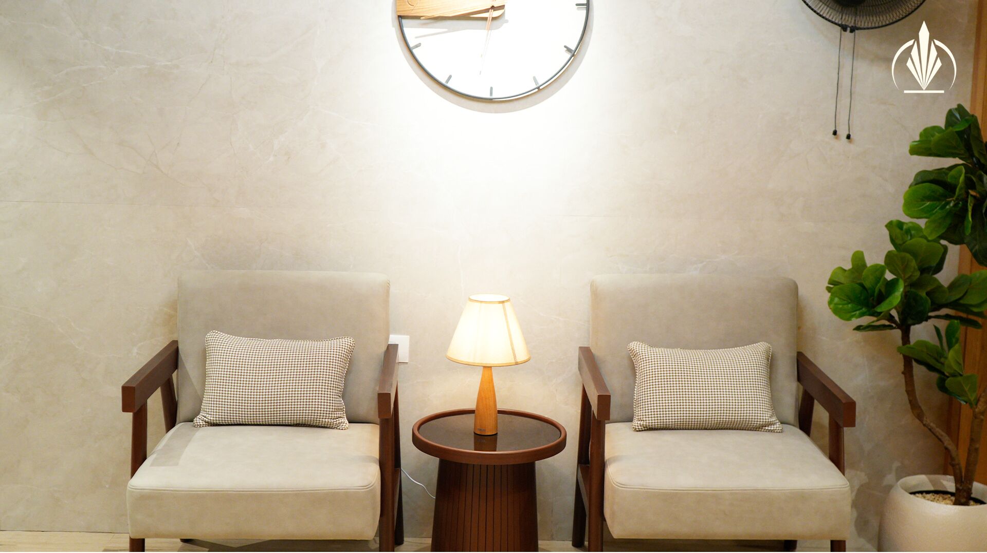Two armchairs with checkered pillows, a round wooden table with a lamp, and a wall clock.