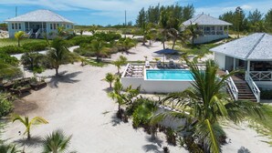 A beachfront property with a swimming pool, multiple beach houses, and lush vegetation. - Brand New Villa on Pink-Sand Beachfront Estate with Shared Pool (Governors Harbour)