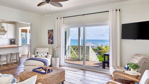 A spacious living room with a large sliding glass door leading to a balcony with a view of the ocean. There is a comfortable seating area with a sofa and armchairs, a coffee table, and a television mounted on the wall. - Brand New Villa on Pink-Sand Beachfront Estate with Shared Pool (Governors Harbour)