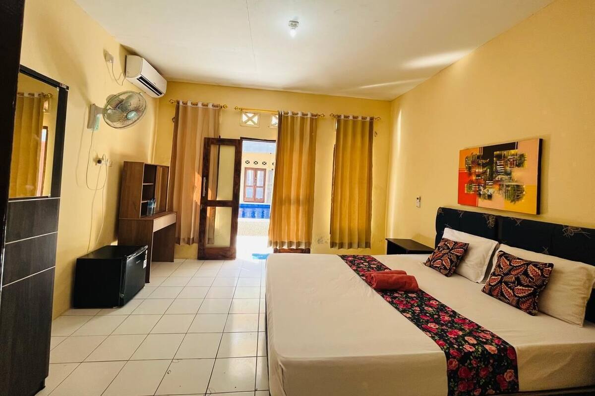 Een hotelkamer met een bed, een bureau, een stoel, een airconditioner, een ventilator en een raam met gordijnen.