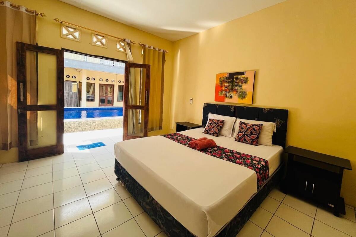 Hotelkamer met een bed, nachtkastje, schilderij en uitzicht op een zwembad en gebouw door een geopende deur.