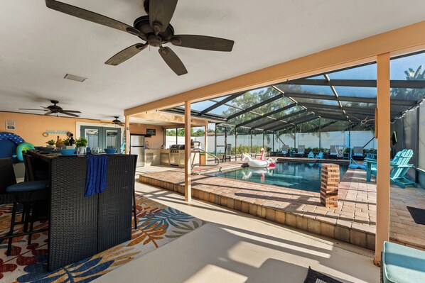A pool area with a covered seating area, a grill, and a ceiling fan.