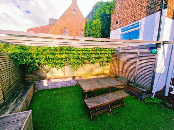 A backyard with a wooden table and benches, a brick wall, and a green hedge.