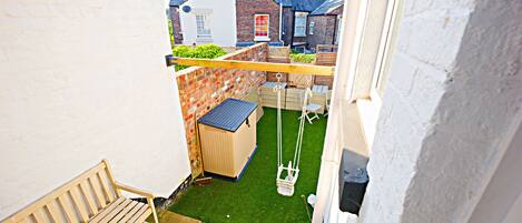 A narrow outdoor space with a wooden bench, a swing set, and a small shed.