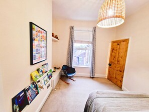 A bedroom with a bed, a desk with books, a chair, a window with curtains, and a wooden door.