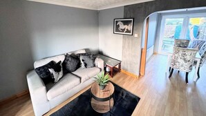 A modern living room with a sofa, a coffee table, and a chair. There is a window with a view of the outside. - Ashford Blissful Retreat Modern 3-bed House (Ashford)