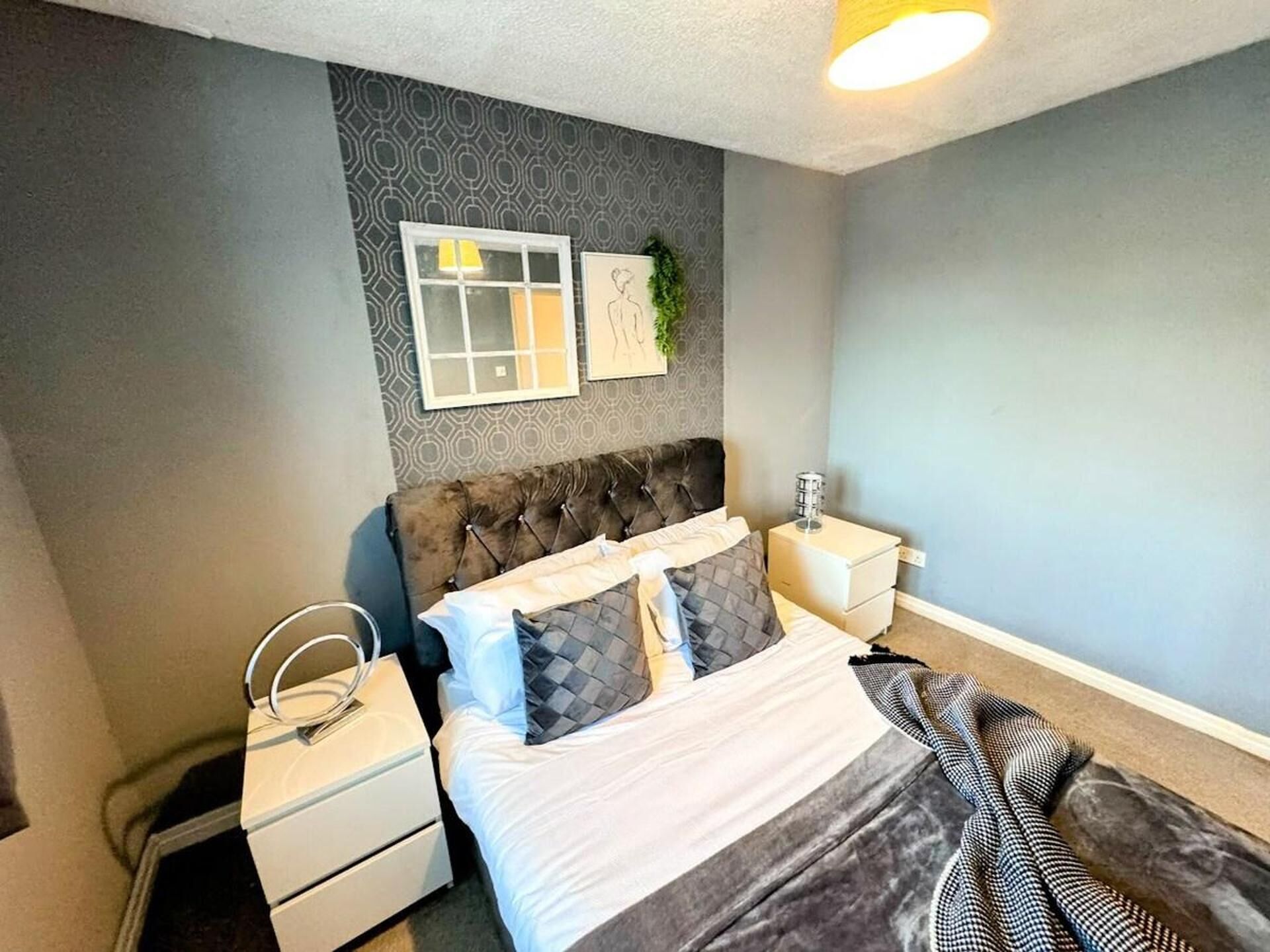 A bedroom with a tufted headboard, a bed with a white and gray striped blanket, two bedside tables with lamps, and a wall-mounted artwork.