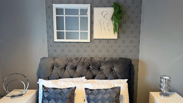 A bedroom with a tufted headboard, a bed with white and dark blue quilted pillows, a white bedside table with a ring light, a framed drawing on the wall, and a small potted plant. - Ashford Blissful Retreat Modern 3-bed House (Ashford)