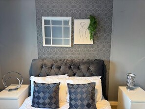 Un dormitorio con cabecera capitonada, cama con almohadones acolchados blanco y azul marino, mesita de noche blanca con aro de luz, un dibujo enmarcado en la pared y una pequeña planta en maceta.