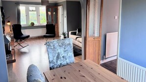 A modern living room with a wooden table, two black chairs, a blue sofa, and a large window providing natural light. - Ashford Blissful Retreat Modern 3-bed House (Ashford)